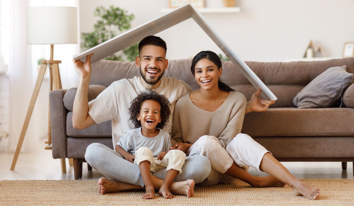 Cheerful parents with child smiling and keeping roof mockup over heads while sitting on floor in cozy living room