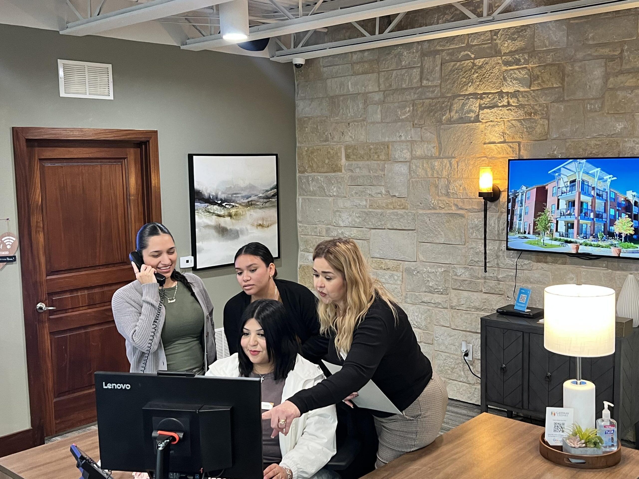 Four women collaborate around a computer in an office; one is on the phone, others are focused on the screen.